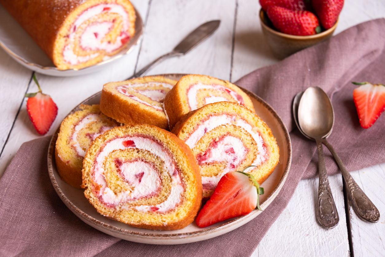 Teller mit mehreren Scheiben einer goldbraunen Biskuitrolle, gefüllt mit Mascarponecreme und Erdbeerstückchen, daneben frische Erdbeeren.