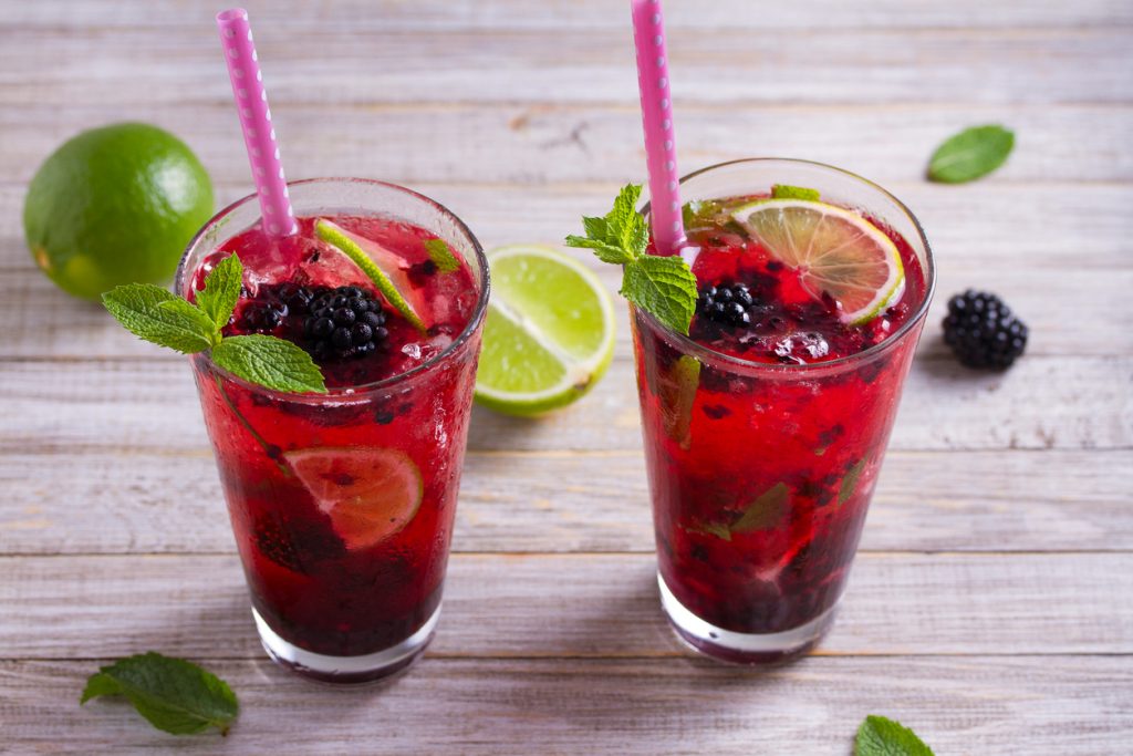 Zwei Gläser Brombeer-Mojito mit Limettenscheiben, Minze und rosa Strohhalmen auf Holz.