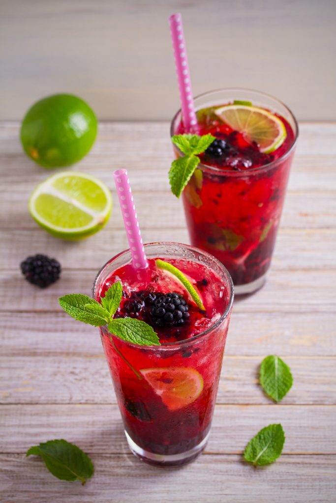 Zwei Gläser Brombeer-Mojito mit Limettenscheiben, Minze, Eis und rosa Strohhalmen.