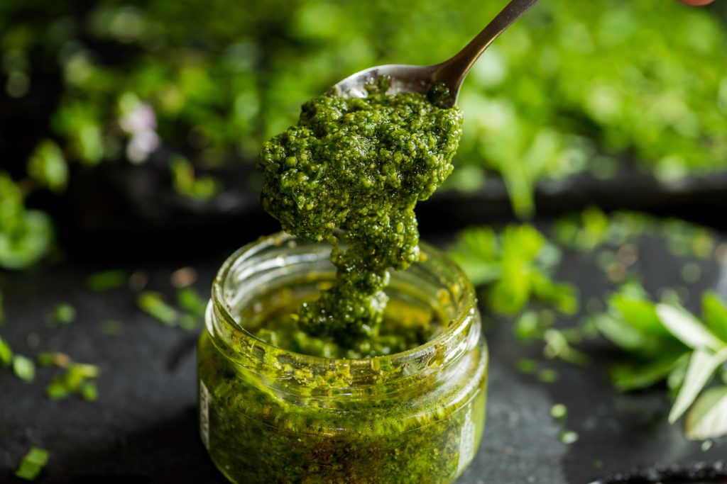 Ein Löffel hebt grünes Petersilienpesto aus einem kleinen Glas auf dunklem Untergrund.
