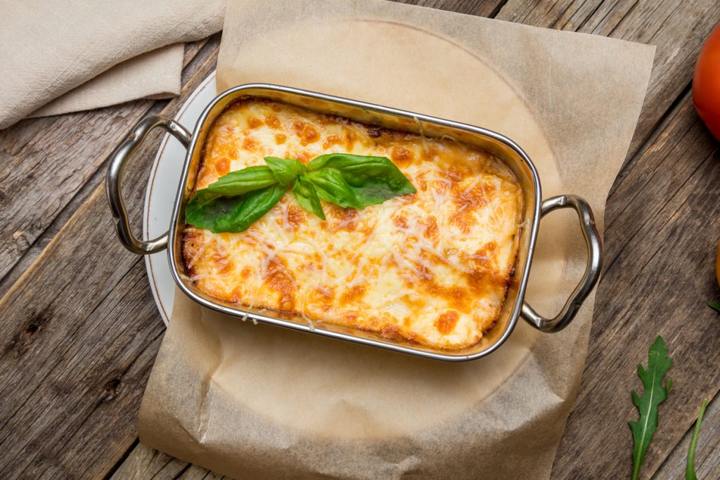 Obersicht auf eine kleine rechteckige Lasagne in Metallform mit geschmolzenem Käse und Basilikum.