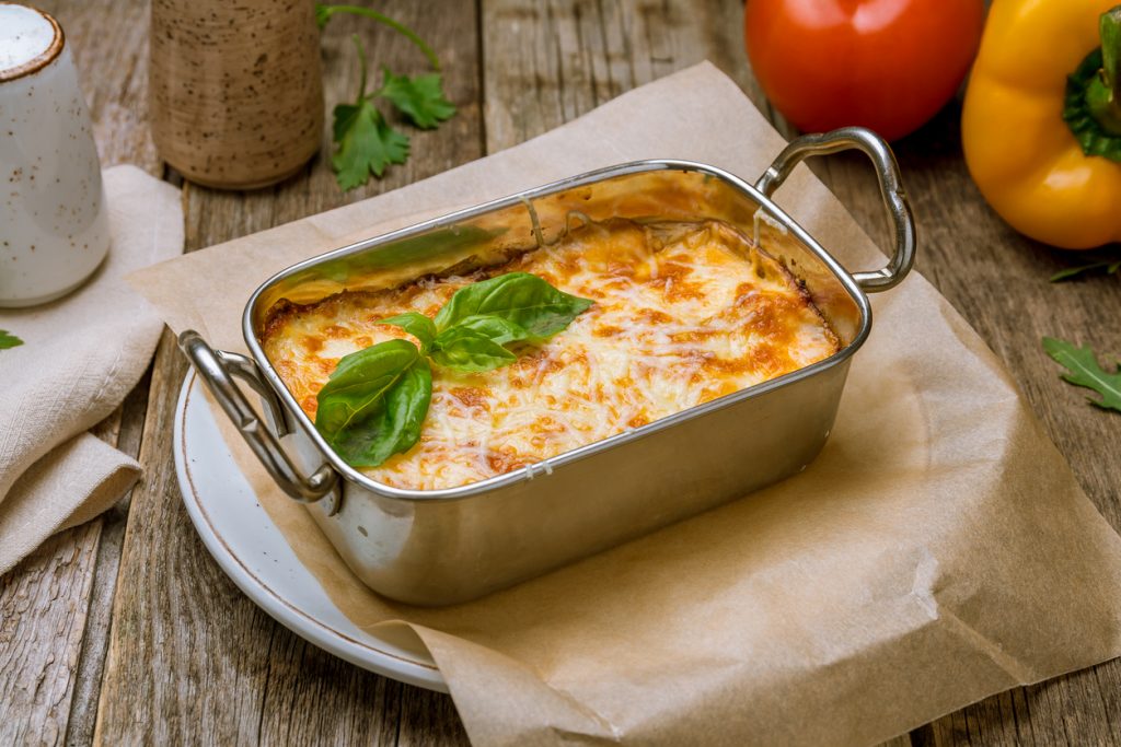 Kleine metallene Auflaufform mit goldbraun überbackener Lasagne und zwei Basilikumblättern auf Holzbrett.