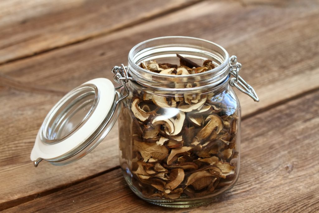 Offenes Bügelglas mit vielen getrockneten Pilzscheiben auf rustikalem Holztisch.