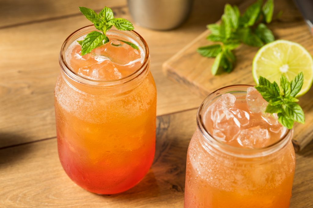 Zwei orange-rote Planters-Punch-Cocktails mit Eis und Minze auf einem Holztisch.