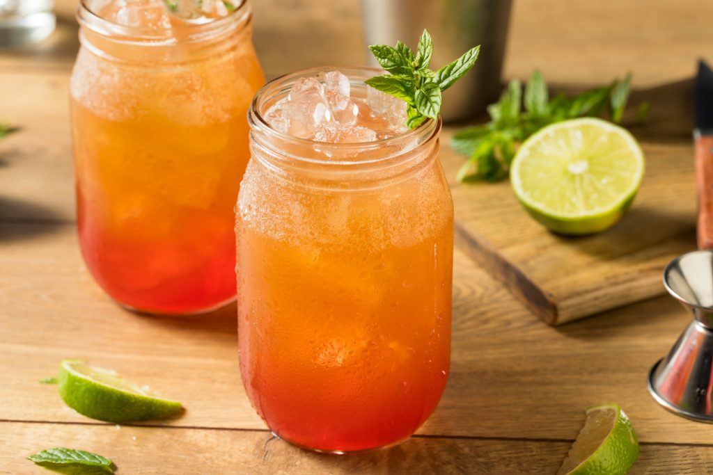 Nahaufnahme eines Planters Punch im Glas mit Eiswürfeln, Minzzweig und Limettenstücken daneben.