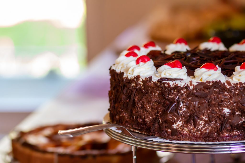 Ganze Schwarzwälder Kirschtorte mit Sahnerosetten, Kirschen und Schokoraspeln auf einem Tortenständer.