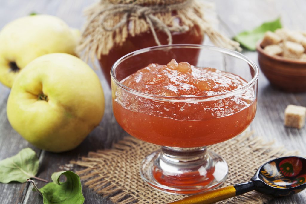 Glas-Schale mit orangefarbener Quittenkonfitüre auf Holztisch, daneben Quitten, Zuckerwürfel und ein dekorativer Löffel.