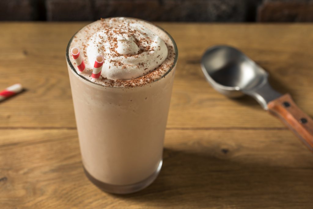 Glas mit Schokoshake, Sahne, Schokoraspeln und zwei rot-weißen Strohhalmen auf Holztisch.