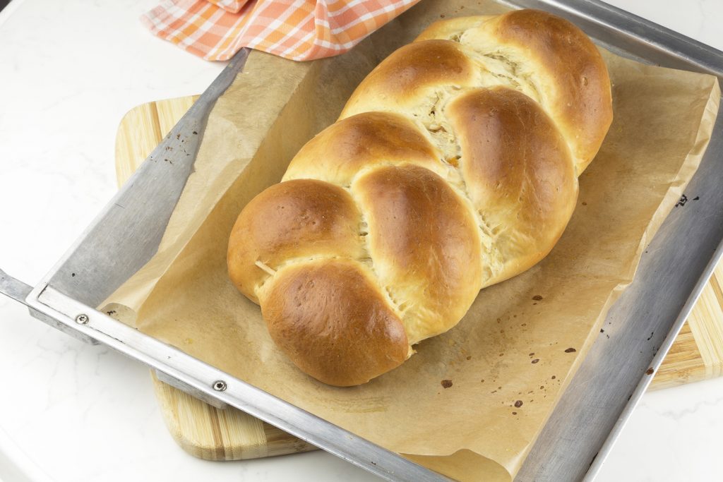 Ein goldbrauner Hefezopf liegt auf Backpapier in einem Metallblech und wirkt hoch aufgegangen.