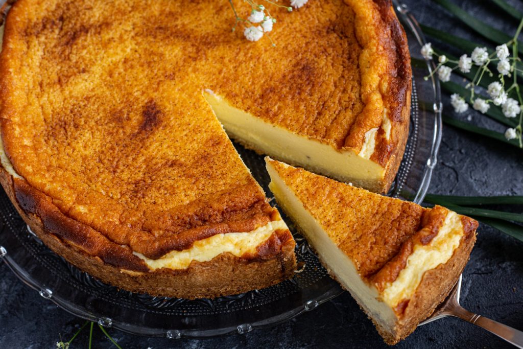 Anschnitt eines klassischen Käsekuchens mit goldbrauner Oberfläche auf Glasplatte, ein Stück wird herausgehoben.