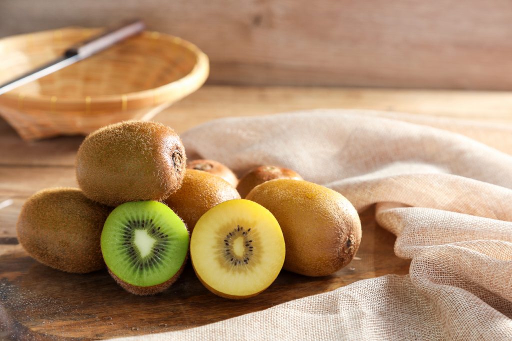 Ganze Kiwis liegen auf Holz, davor halbierte grüne und gelbe Früchte.