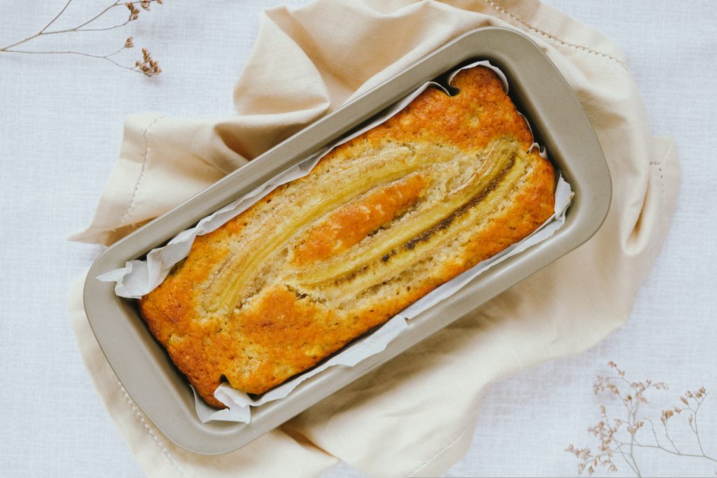 Goldbraunes Bananenbrot mit längs aufgelegter Banane in Backpapier in einer Kastenform, von oben fotografiert.