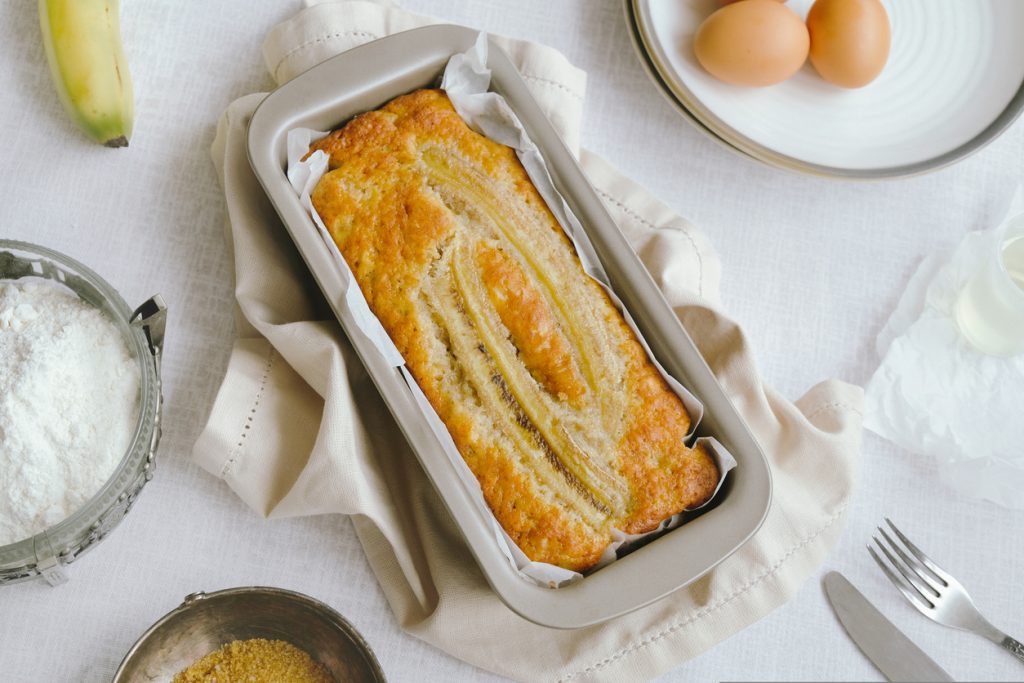 Frisch gebackenes Bananenbrot mit längs aufgelegter Banane in der Kastenform, umgeben von Eiern, Mehl und Zucker.