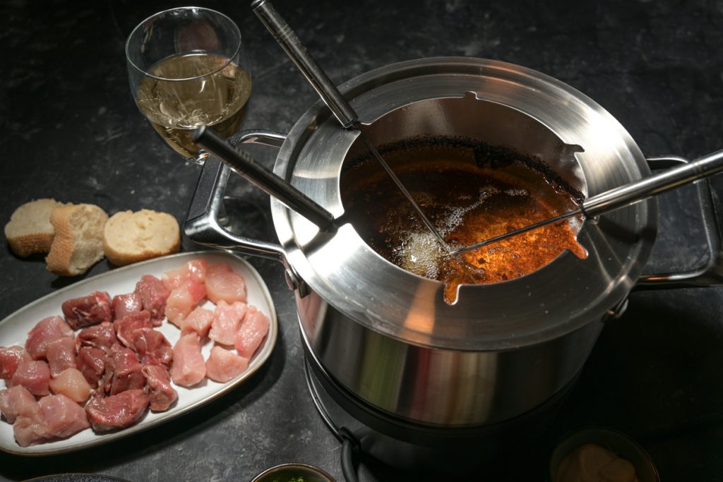 Ein heißer Fonduetopf mit Öl steht neben Fleischwürfeln, Brot und einem Weinglas.