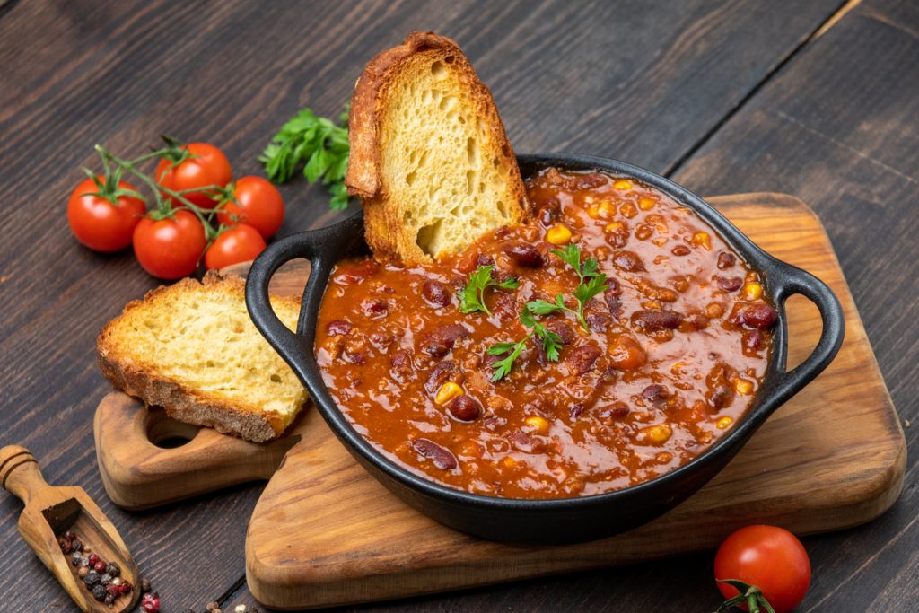 Gusseisentopf mit Chili, Toastscheibe und Petersilie auf einem Holzbrett.