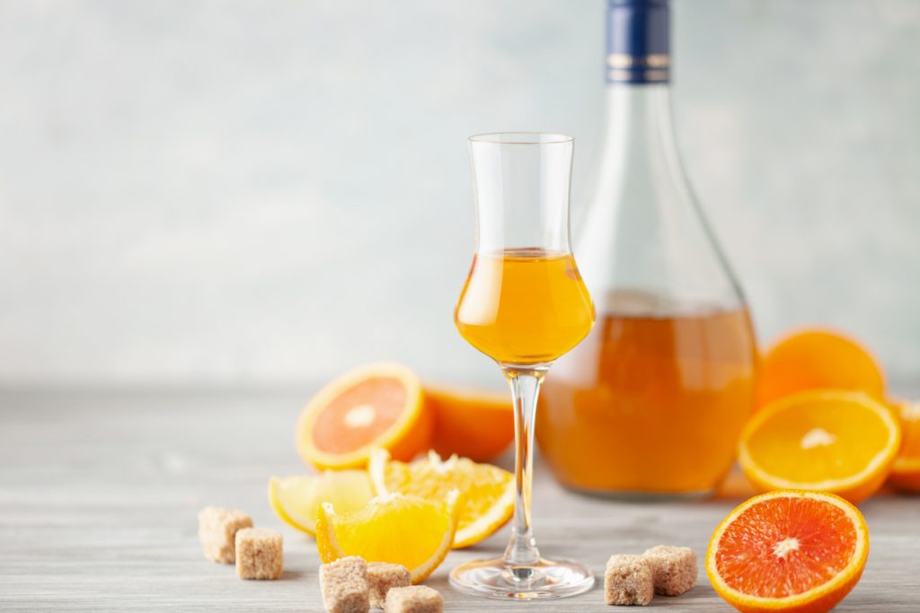 Ein schlankes Likörglas mit Orangenlikör steht vor einer Flasche, daneben liegen Orangenhälften und braune Zuckerwürfel.