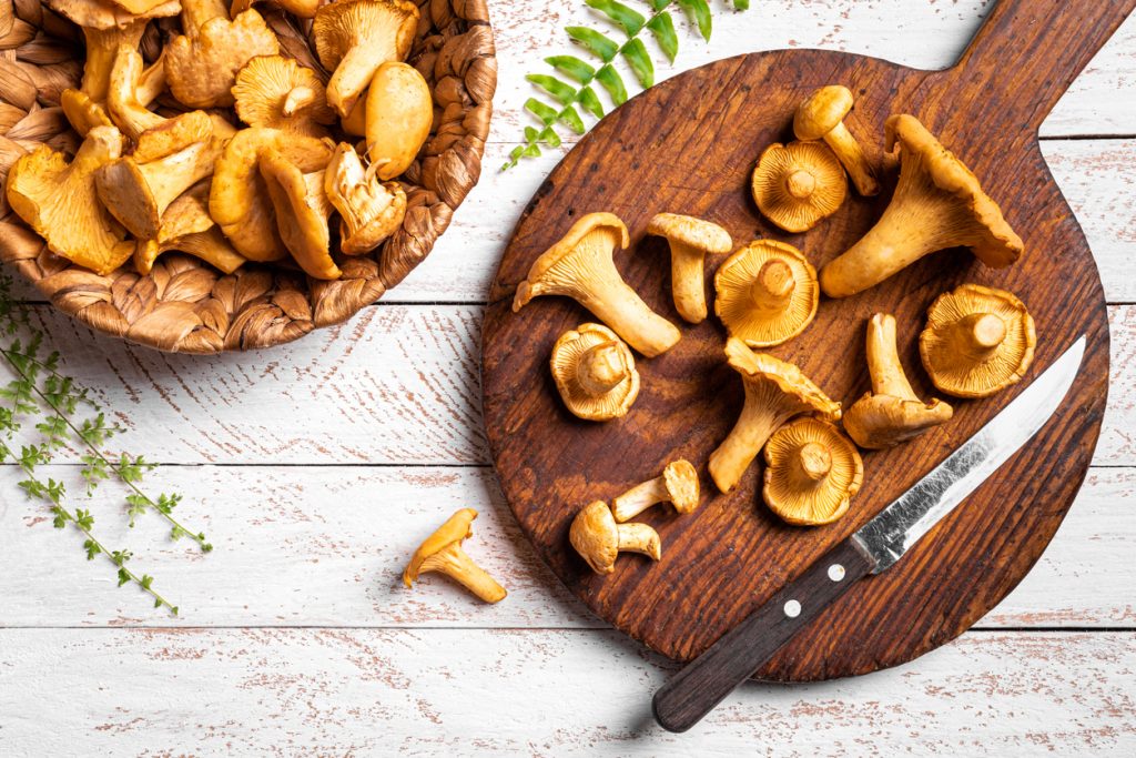 Frische Pfifferlinge auf rundem Holzbrett mit Messer, daneben ein Korb auf weißem Holztisch.