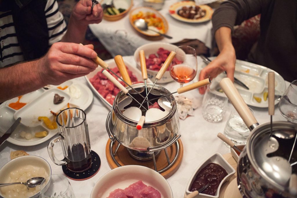 Hände tauchen Fonduegabeln in einen Edelstahltopf, umgeben von Fleisch, Dips und Getränken.