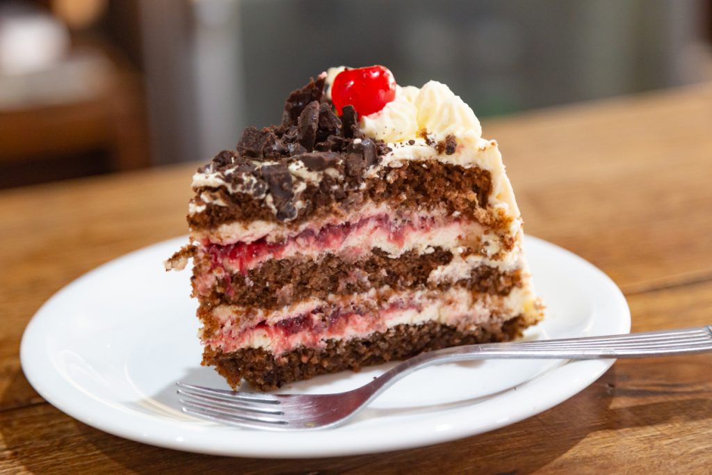 Ein Stück Schwarzwälder Kirschtorte mit Sahne, Kirschen und Schokoraspeln auf weißem Teller.