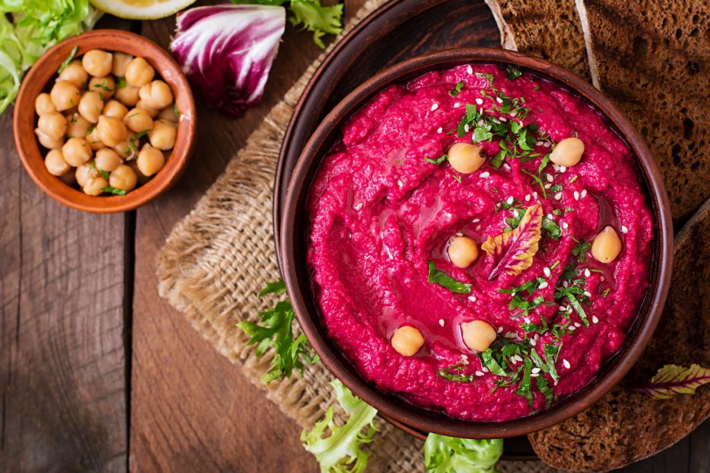 Obenansicht einer Schale pinken Hummus mit Kichererbsen, Kräutern und Sesam, flankiert von Brotscheiben.
