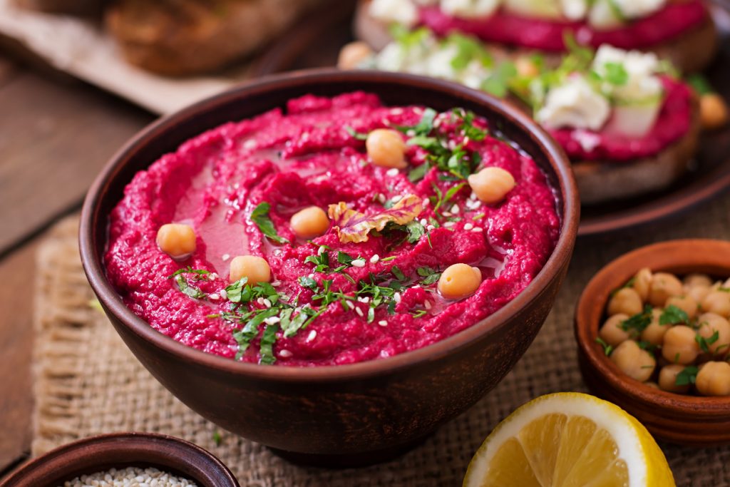 Schale mit pinkem Rote-Bete-Hummus, Kichererbsen, Kräutern und etwas Öl, daneben Zitrone.