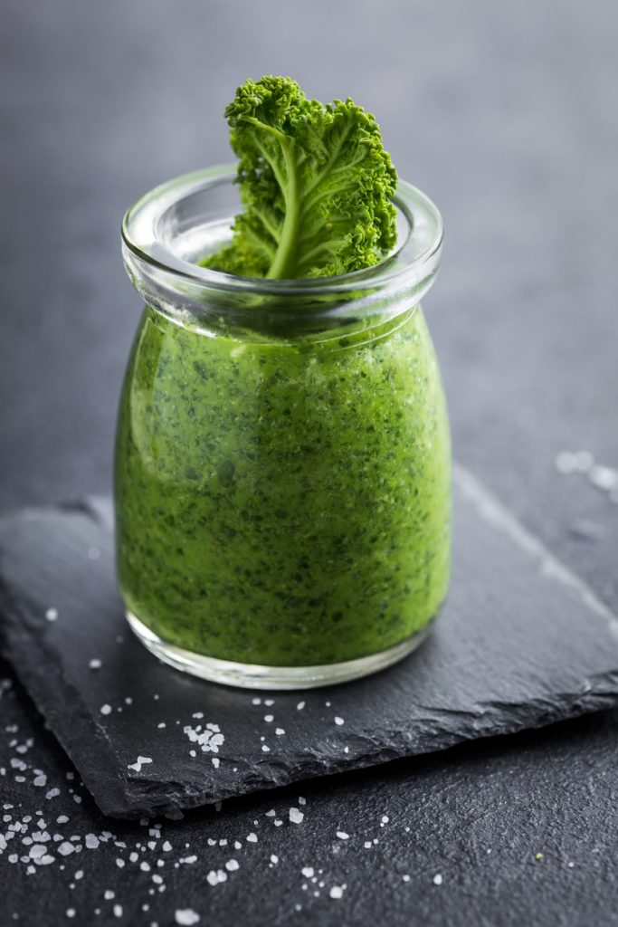 Glas mit leuchtend grünem Pesto, oben steckt ein Grünkohlblatt auf einer Schieferplatte mit Meersalz.