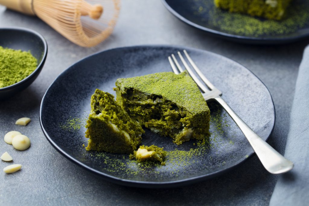 Anschnitt eines grünen Matcha-Brownies mit weißen Schokostücken auf schwarzem Teller neben einer Gabel.