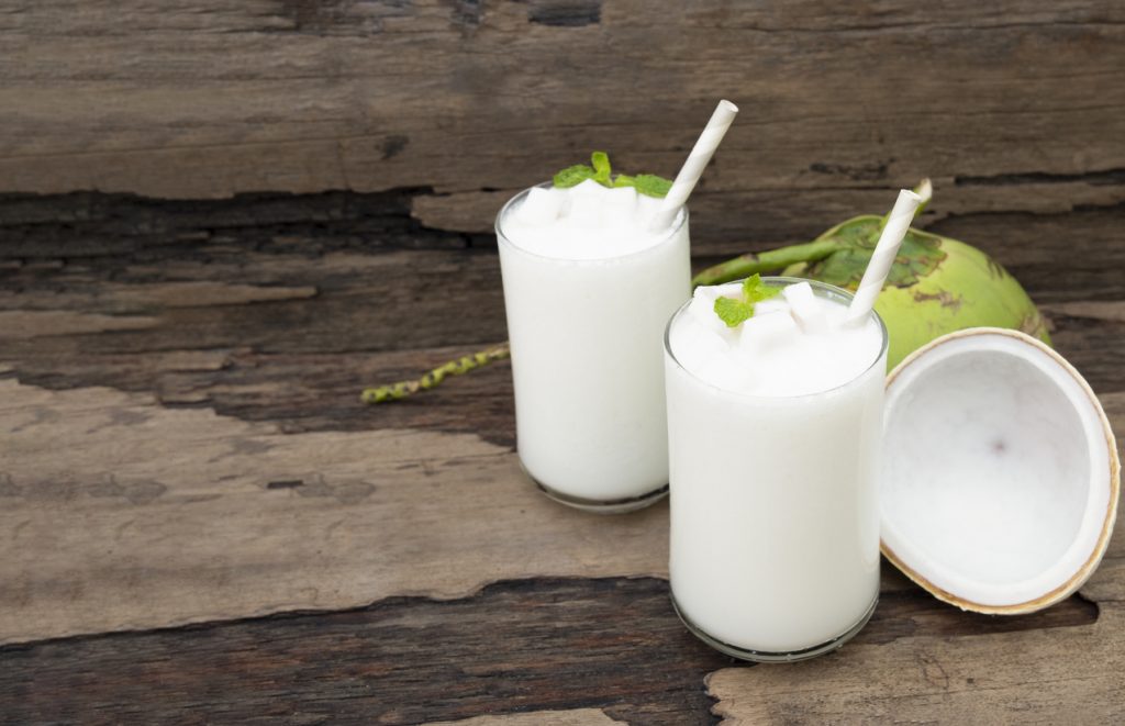 Zwei Gläser Kokos-Milchshake mit Strohhalm auf Holz, garniert mit Minze und Kokosstücken.