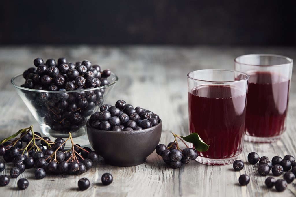 Schalen mit dunklen Beeren und zwei Gläser mit violettem Saft auf einem Holztisch.