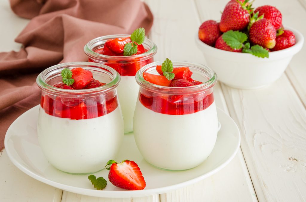Drei Gläser Panna Cotta mit Erdbeersauce und Minze stehen auf einem weißen Teller neben einer Schale Erdbeeren.