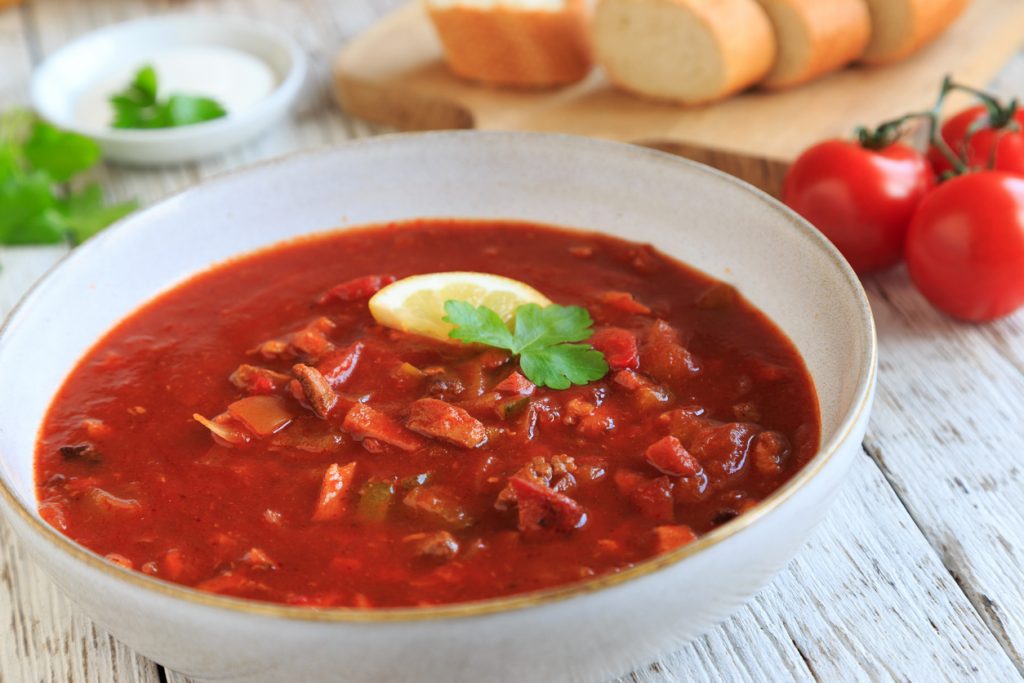 Teller mit roter Soljanka, Zitronenscheibe und Petersilie, daneben Baguette.