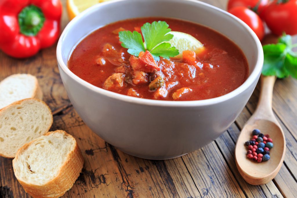 Schüssel mit roter Soljanka, Petersilienblatt und Zitronenspalte, Brot und Gewürze daneben.