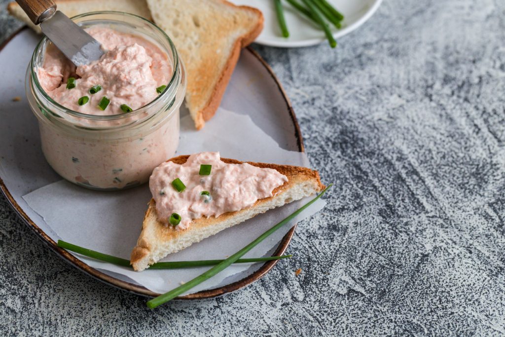 Dreieckstoast mit rosa Lachsfrischkäse und Schnittlauch neben einem Glas.