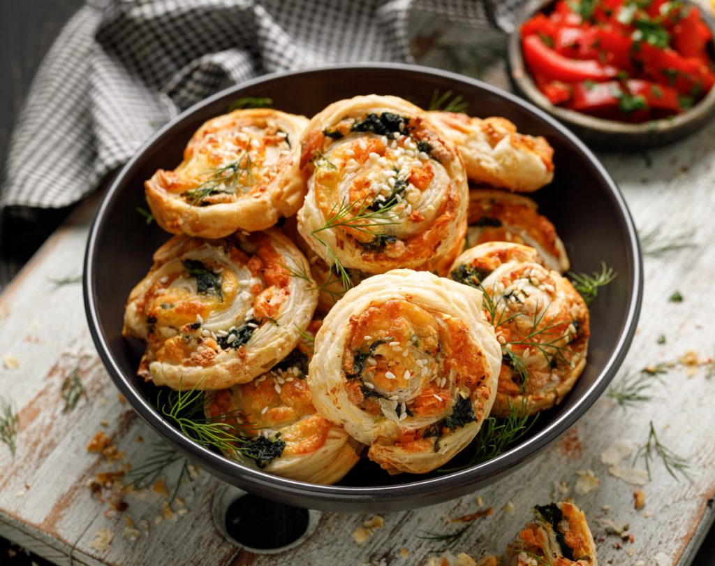 Eine Schüssel voller goldener Blätterteig-Pinwheels mit Lachs, Spinat und Käse, garniert mit Dill.