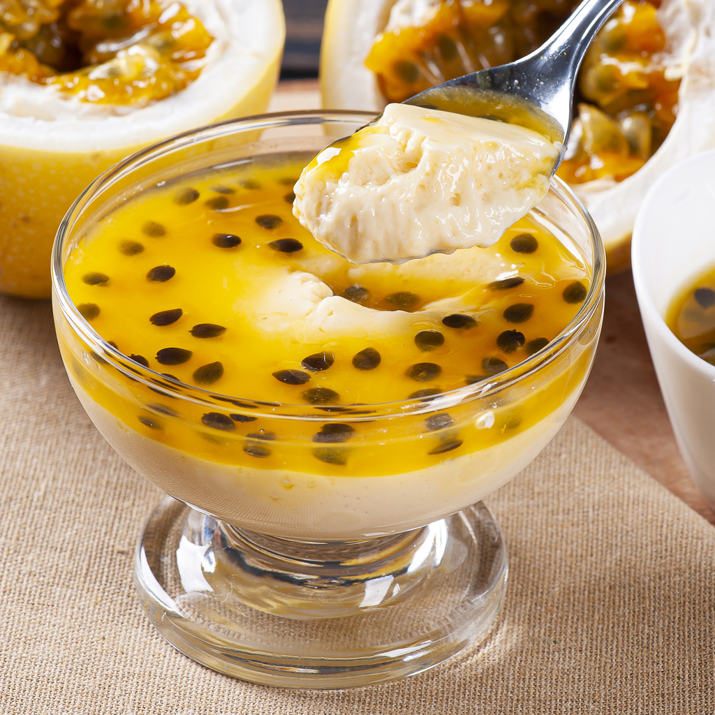 Nahaufnahme einer Dessertschale mit gelbem Passionsfruchtspiegel, in den ein Löffel sticht.