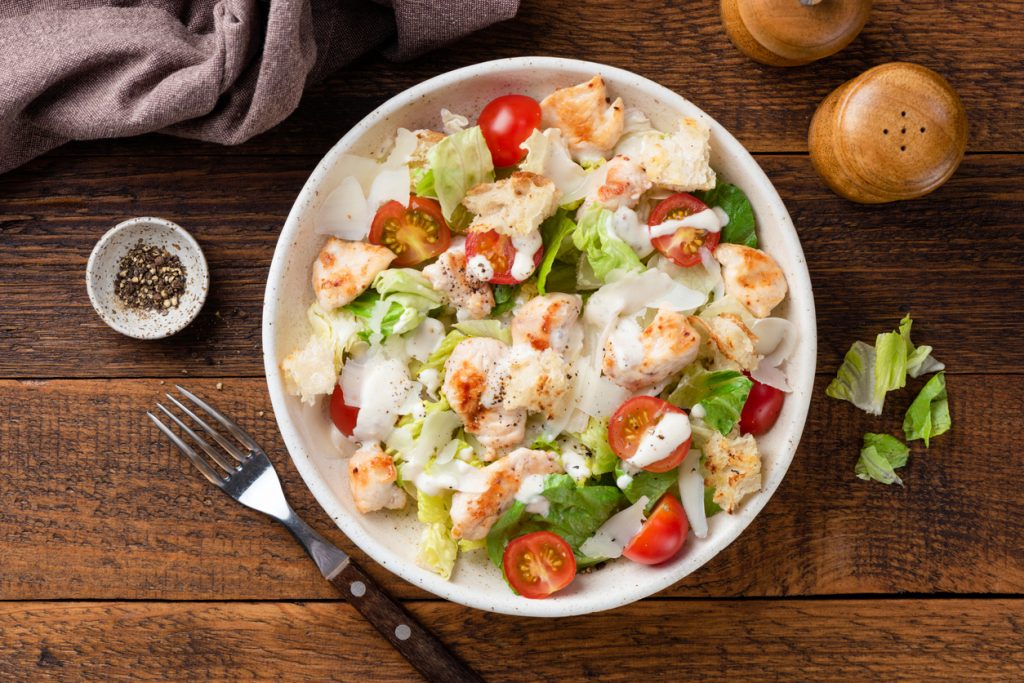 Schüssel mit Caesar Salad aus Römersalat, Hähnchen, Croûtons, Kirschtomaten und Parmesanspänen von oben.