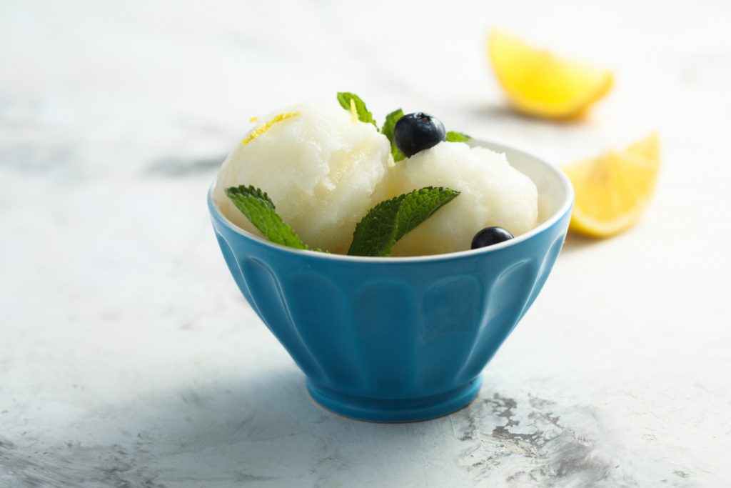 Zwei Kugeln Zitronensorbet mit Minze und Blaubeeren in blauer Schale, hinten Zitronenspalten.