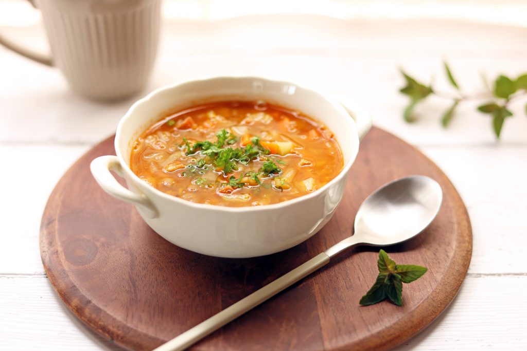 Weiße Suppenschale mit rot-orangener Linsen-Gemüse-Suppe und Petersilie auf einem runden Holzbrett mit Löffel.