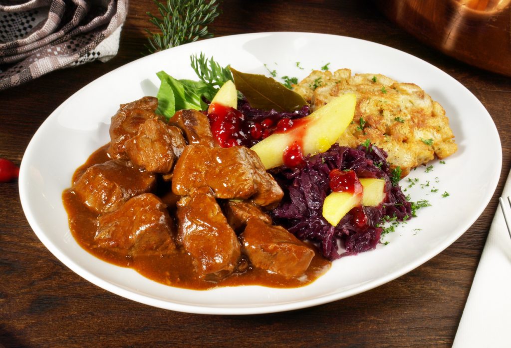 Teller mit Hirschragout in dunkler Sauce, Rotkohl, Apfelspalten mit Preiselbeeren und gebratener Knödelscheibe.