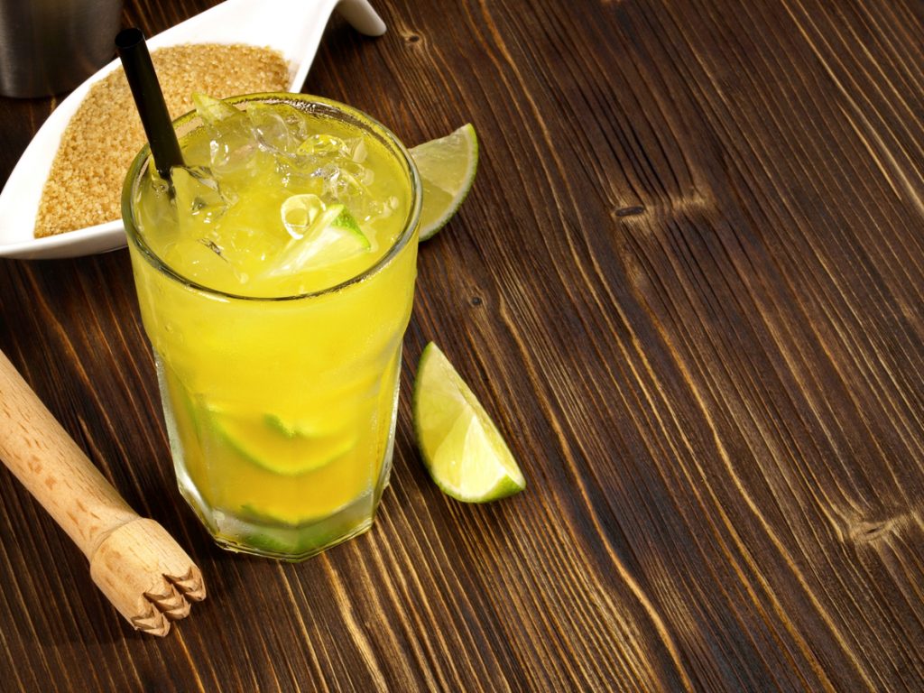 Gelber Ipanema-Mocktail im Glas mit Eiswürfeln, Limettenspalten und Strohhalm auf Holztisch.