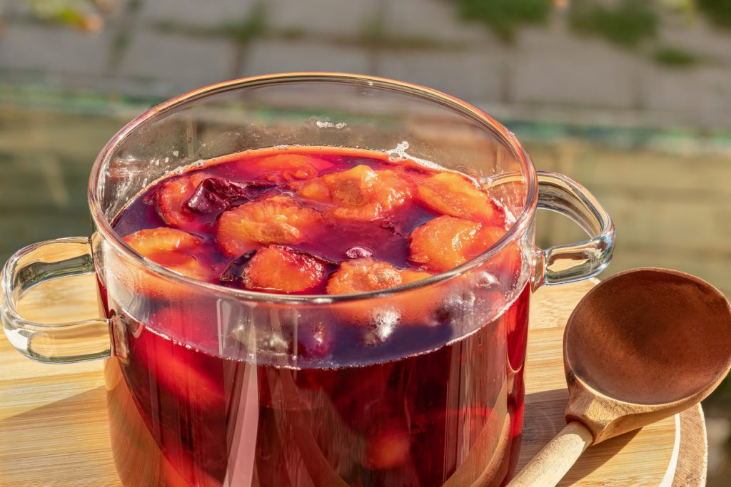 Durchsichtiger Topf mit rotem Kompott und Pflaumenstücken auf Holzbrett.