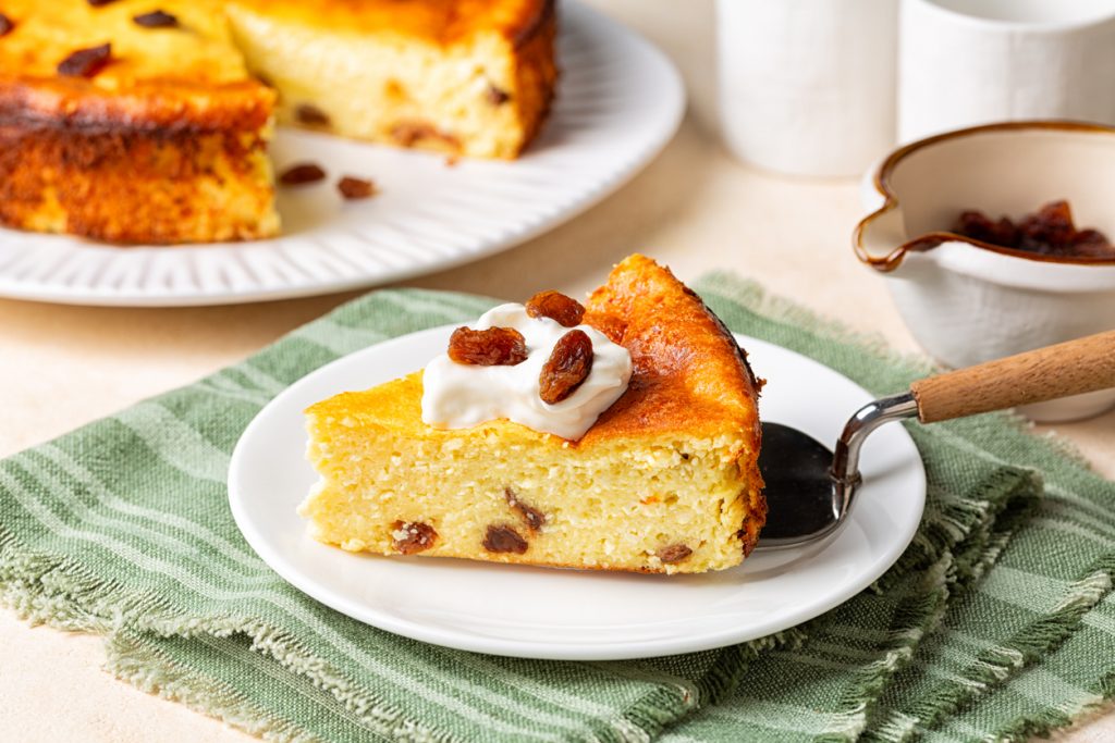 Stück Käsekuchen mit Rosinen und Sahnehaube auf einem Teller vor dem restlichen Kuchen.