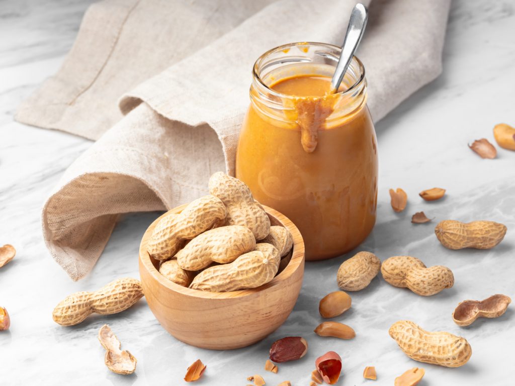 Glas Erdnussbutter mit Löffel neben Schälchen voller Erdnüsse und Schalen.