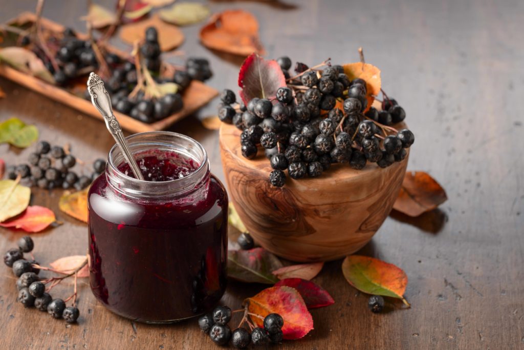 Ein Glas mit dunkler Beerenmarmelade steht neben einer Holzschale mit Beeren und Herbstblättern.
