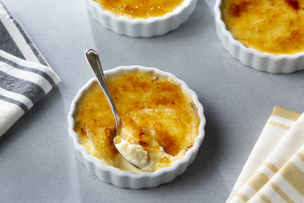 Weiße Crème-brûlée im Ramekin, ein Löffel bricht die karamellisierte Oberfläche.