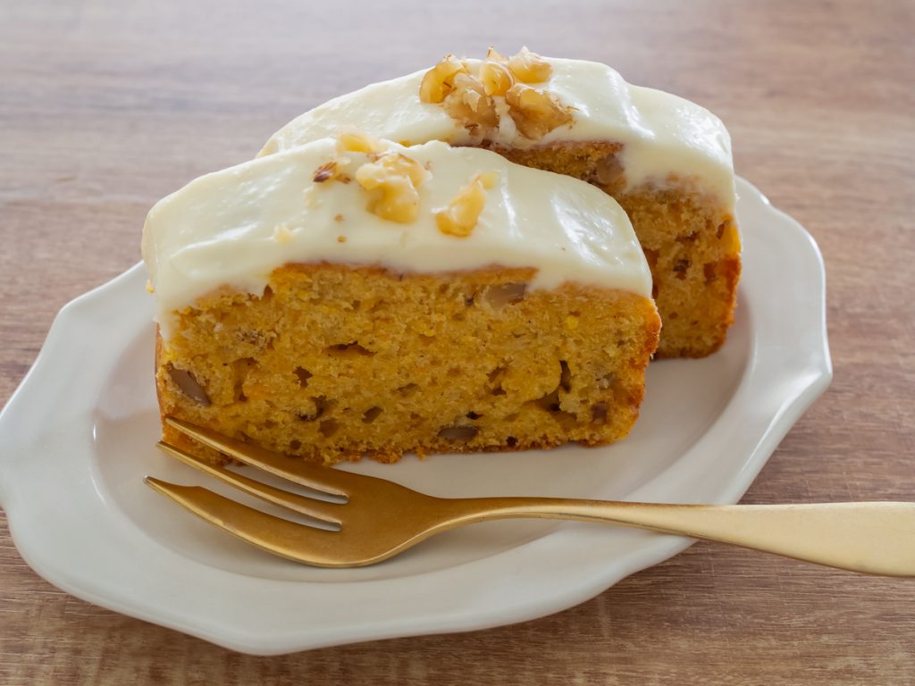 Zwei Stück Karottenkuchen mit dicker Frischkäseglasur und Walnüssen auf einem Teller mit goldener Gabel.