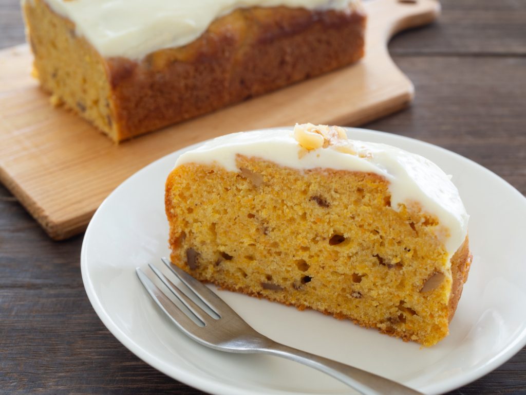 Stück Karottenkuchen mit heller Frischkäseglasur auf einem weißen Teller vor dem restlichen Kuchen.
