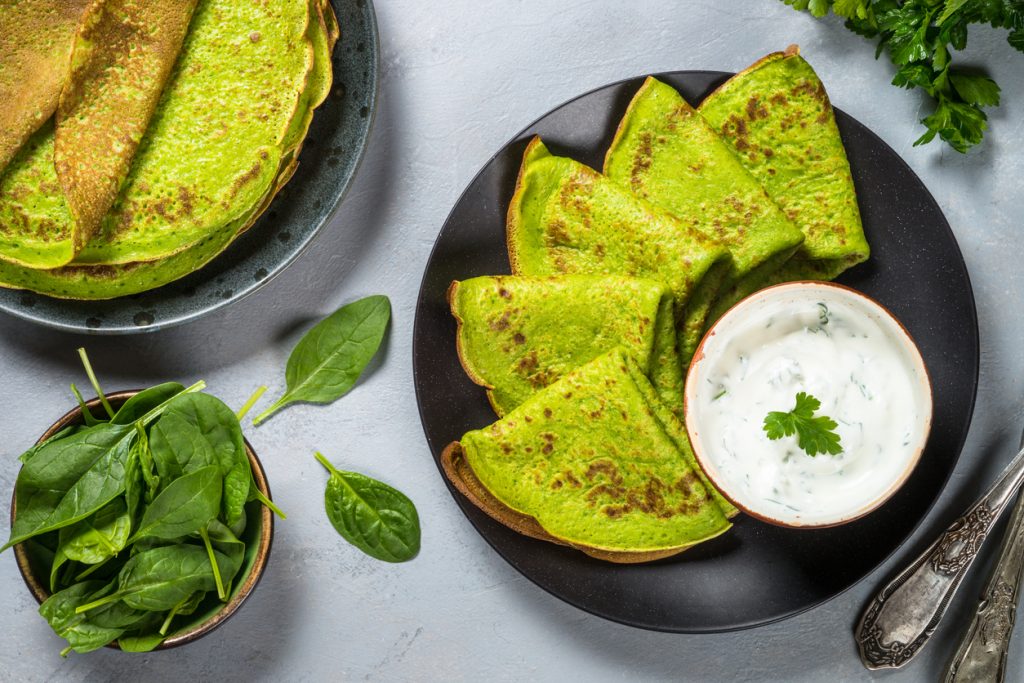 Obenansicht von grünen Pfannkuchen auf zwei Tellern mit Spinatblättern und Schale Dip.