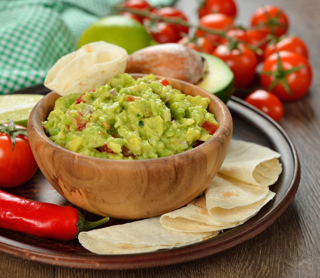 Holzschale mit grob pürierter Guacamole auf einem Teller mit Fladenbrot und Gemüse.