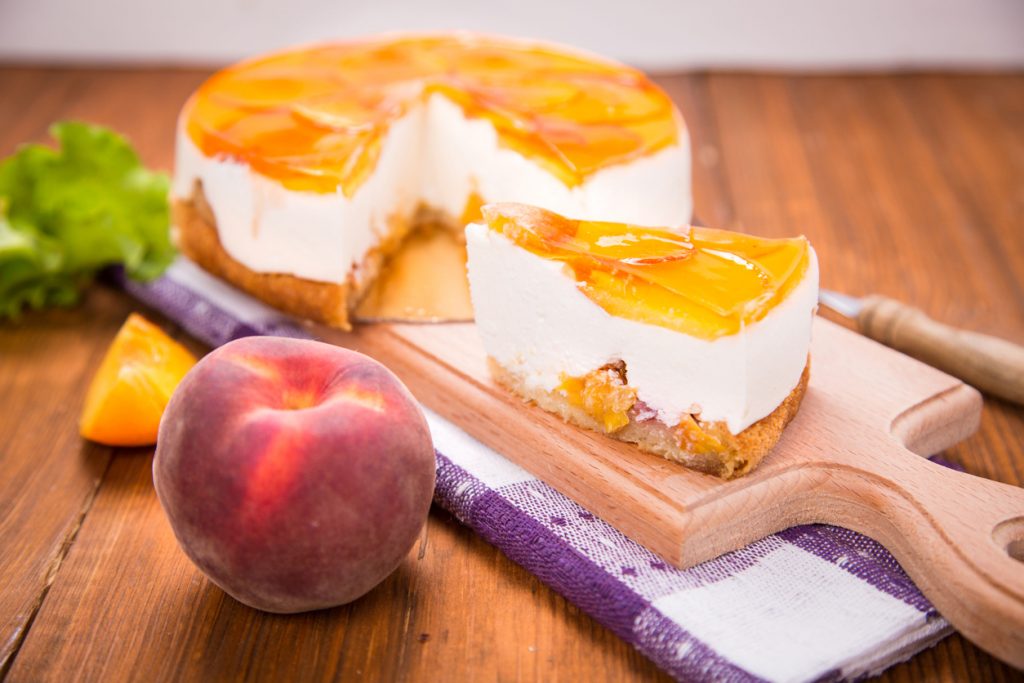 Tortenstück mit Sahneschicht und Pfirsichspiegel auf Holzbrett vor der restlichen Torte.